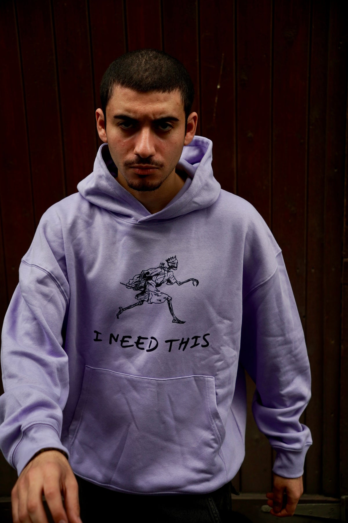 A man wearing a lavender purple hoodie with a black graphic of a skeleton running and the text 'I NEED THIS' printed on the front, posed against a dark brown wooden background.